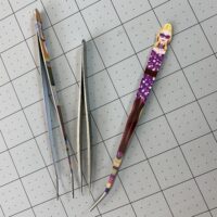 Three metal tweezers lay on top of a gray cutting mat.  Two of the tweezers are painted with people's faces.  