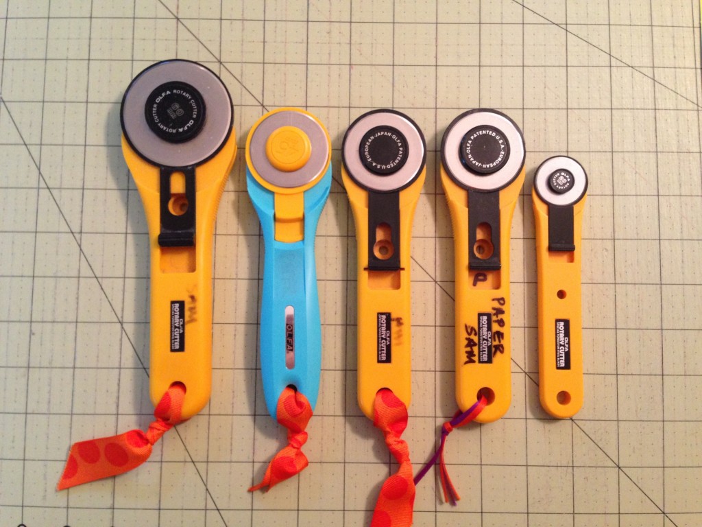 Five rotary cutters are lined up on a cutting surface. Some cutters are written on with permanent markers to indicate what they should be used for. 