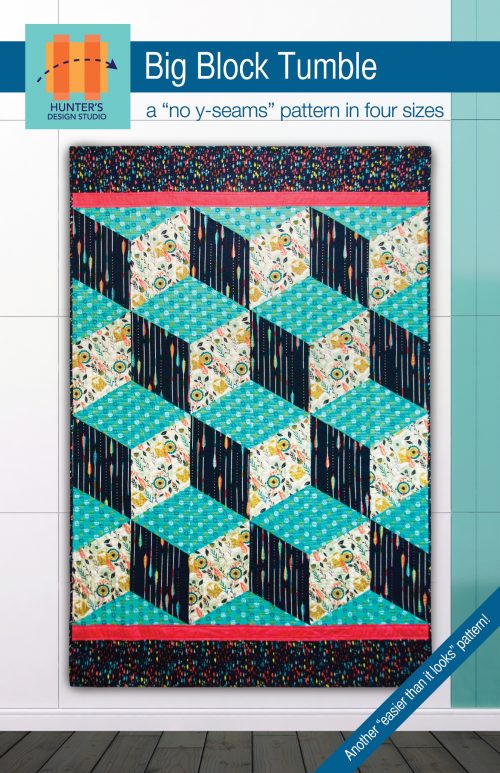 Image is of the Big Block Tumble quilt hanging up on a wall with a floor underneath. The quilt is in the colors of aqua, navy and cream.