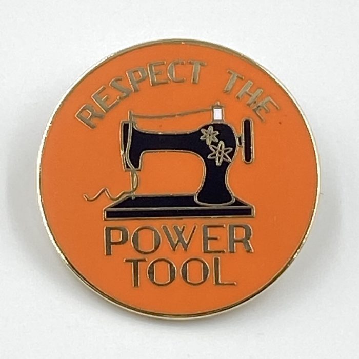 The Respect the Power Tool enamel pin features a black old-fashioned sewing machine with the words 'respect the power tool surrounding it. The background is 'Sam Hunter orange'