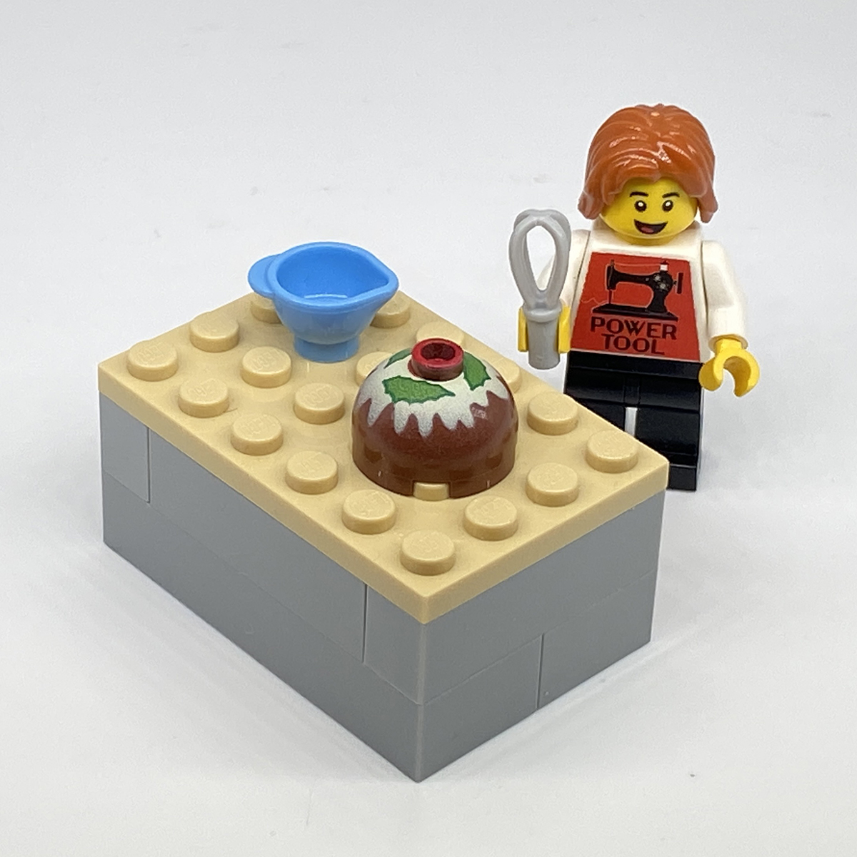 Lego Sam holds a wisk in her right hand and stands behind a lego table with a blue mixing bowl and a finished Christmas Pudding on it