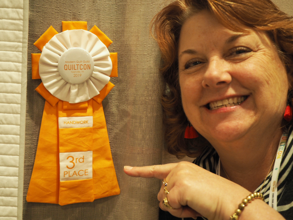 In this image, Sam Hunter is pointed to an orange third place 2019 QuiltCon Ribbon for the Handwork category 
